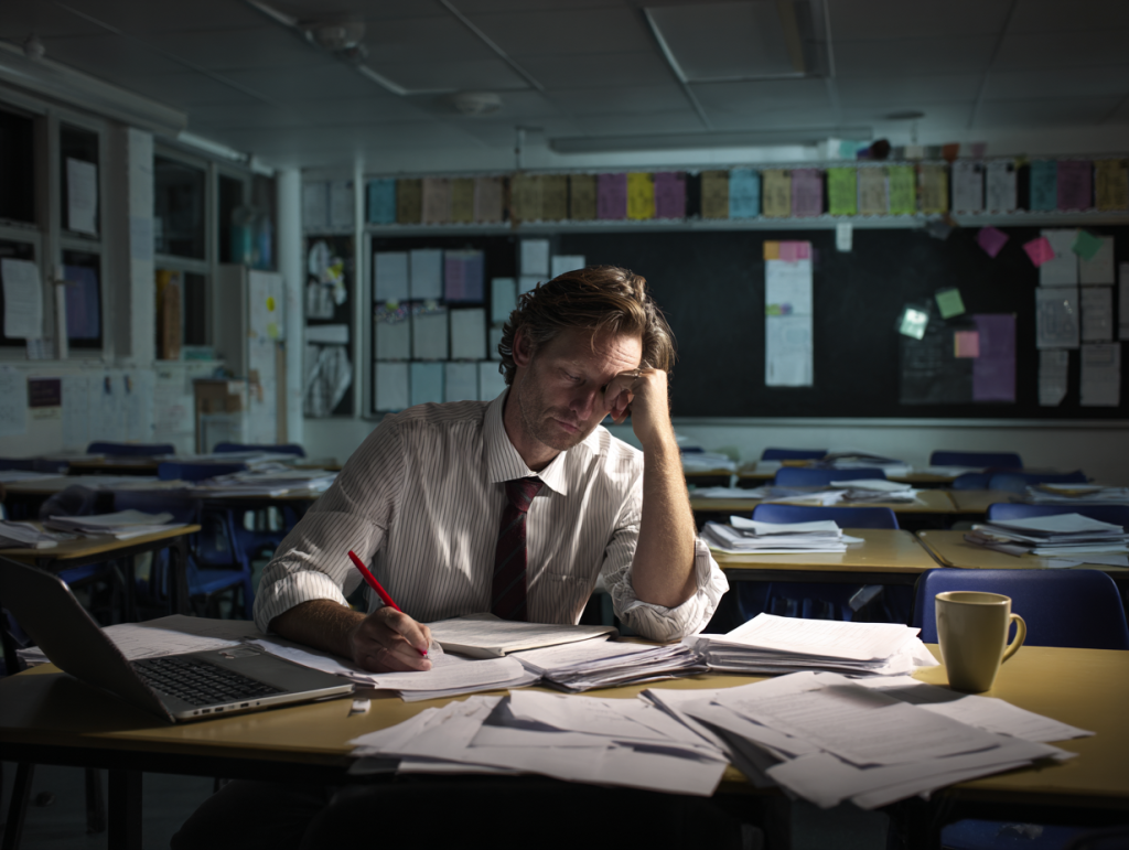 A weary male secondary school teacher sits alone in a UK classroom, cross-referencing papers against a laptop. Surrounded by stacks of documents, he holds a red pen and leans on his hand, his expression tired and resigned. Blue plastic chairs and a cluttered noticeboard frame the scene, lit by a single harsh fluorescent light.